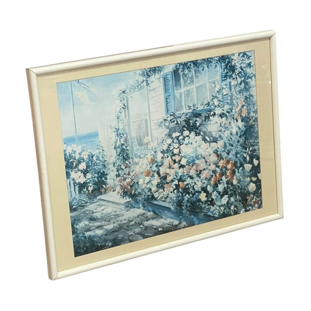 Vintage framed landscape print depicting a coastal cottage surrounded by a densely planted flower garden. The composition...