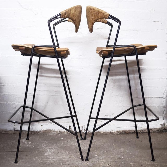 A pair of Arthur Umanoff mid century bar stools, for Raymor. They feature wood slat seats, iron frames with wicker wrapped...