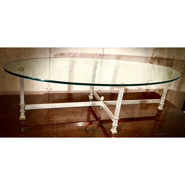 A Maison Jansen style oval iron and brass coffee table with a thick glass top, straight stretchers and a centered acorn...