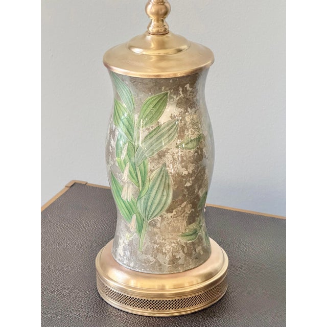 A vase-form glass table lamp decorated with botanical flowers, foliage, and butterflies against a mottled silver-colored...