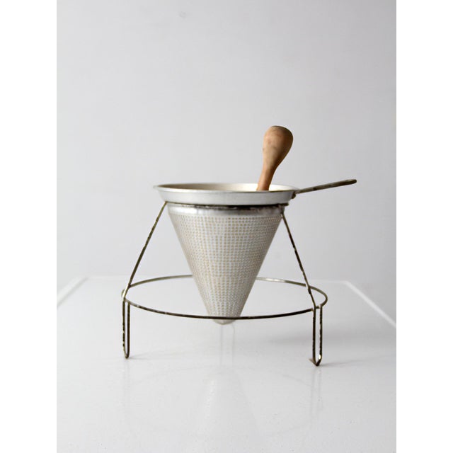 A mid-century conical strainer set with stand and wooden pestle. The perforated metal cone rests in a simple wire frame...