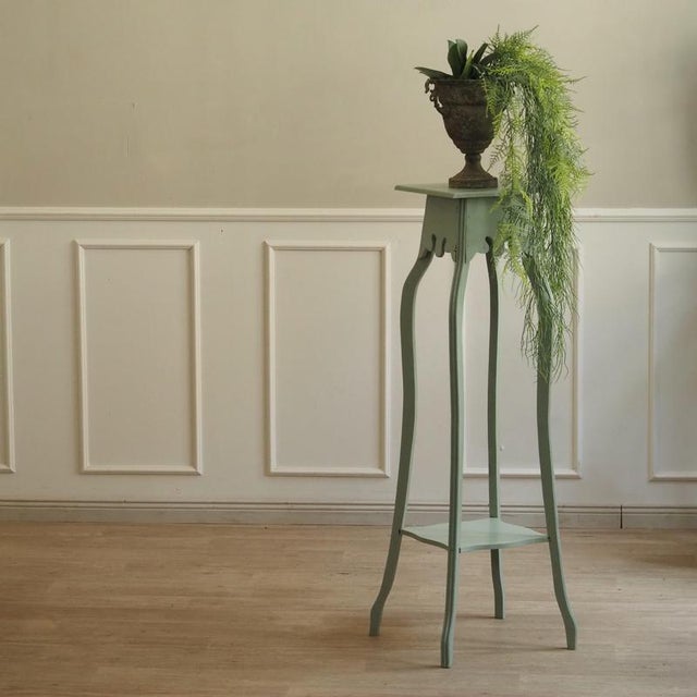 A mint green, lacquered wooden flower stand with two shelves and curved legs. Its design is inspired by art nouveau flower...