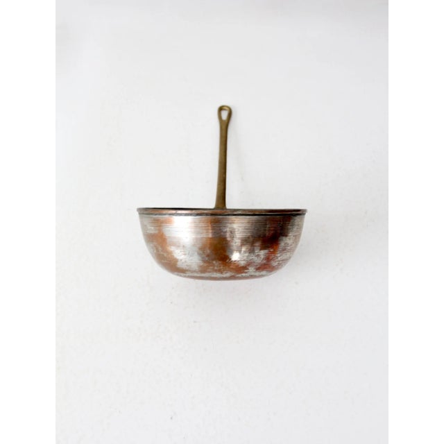 Antique deep-sided copper pan featuring a rounded form with tall walls and a long brass handle pierced with a hanging...