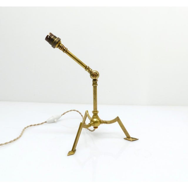 A tripod brass lamp base with an articulated structure, resting on three angled feet characteristic of the Arts & Crafts...