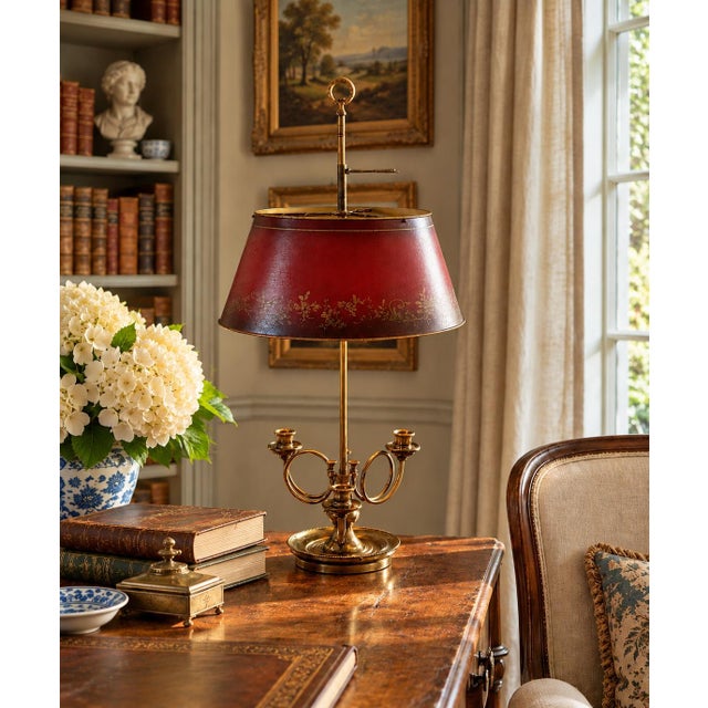 A classic bouillotte lamp rendered in warm brass, distinguished by its sculptural three horn base and tailored red tole...