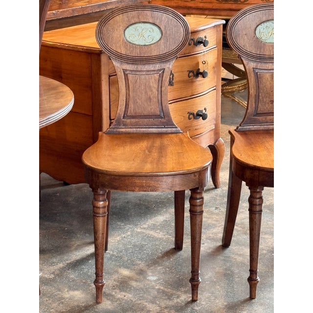 Pair of English hall chairs with shaped seats and oval backs, each centered with a painted and gilt monogrammed oval...