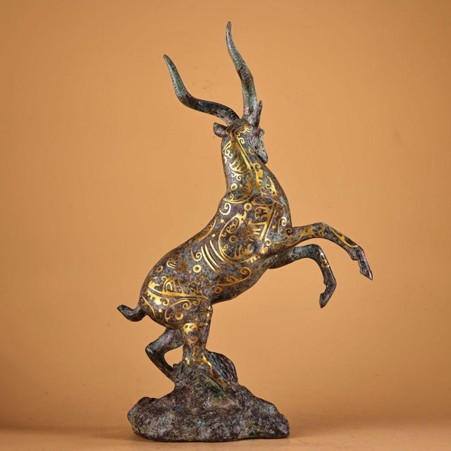 Dynamic archaic-style bronze sculpture of a leaping antelope, posed mid-stride upon a textured rock base. The body is...