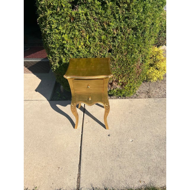 A gold-finished French-style 2-drawer accent table, closely matching models such as the Baxton Studio Patrice 2-Drawer...