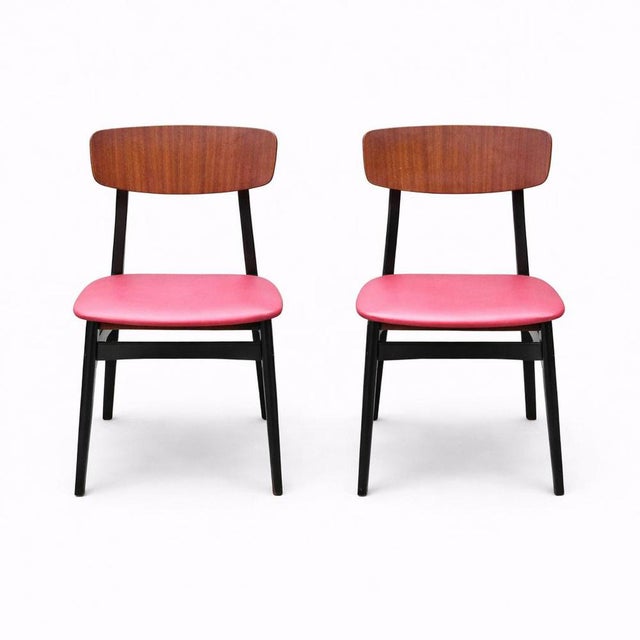 A stylish pair of mid-century dining chairs featuring warm teak backrests, sleek black frames, and vibrant red vinyl...