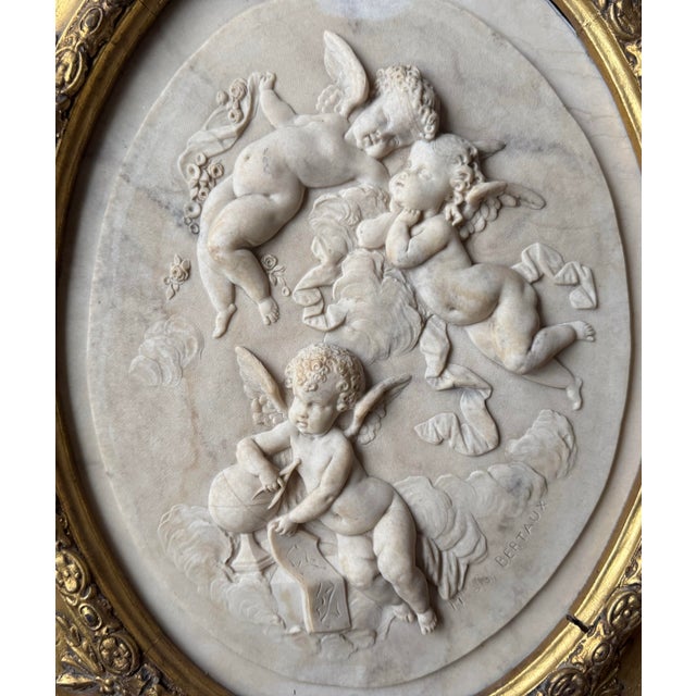 A striking neoclassical carved marble bas-relief depicting three cherubs in high relief, presented in an ornate oval gilt...