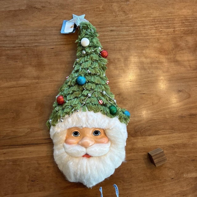 The Katherine's Collection Santa with Tree Hat Wall Mask Hanging is a festive and charming addition to your holiday decor....