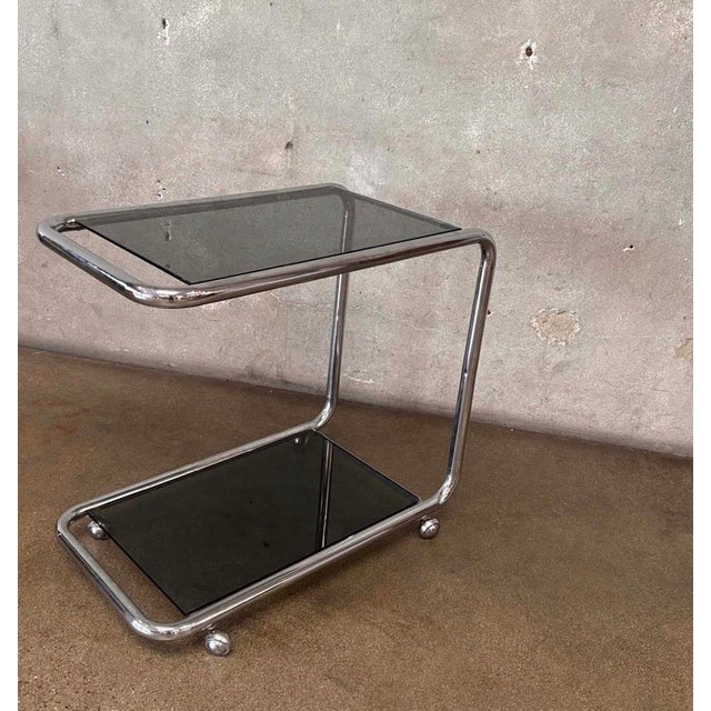 Vintage chrome bar cart featuring a sculptural tubular frame, two smoked-glass shelves, and rolling casters. Its clean...