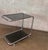 Vintage chrome bar cart featuring a sculptural tubular frame, two smoked-glass shelves, and rolling casters. Its clean...