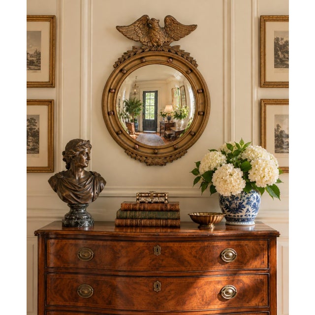 A Federal-style giltwood convex mirror crowned with a carved eagle, its wings outstretched above a laurel motif—an...