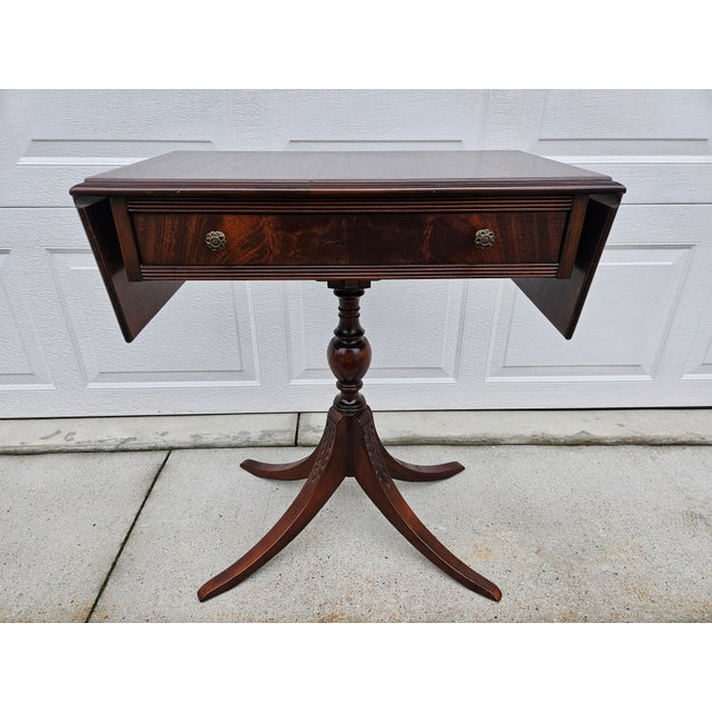 A traditional mahogany drop leaf side table with a carved pedestal base. This piece has one drawer with decorative brass...