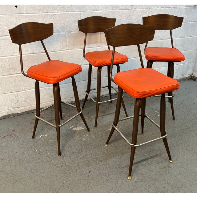 This is a vintage mid-century modern swivel bar stool featuring a bright orange vinyl seat and a curved wooden laminate...