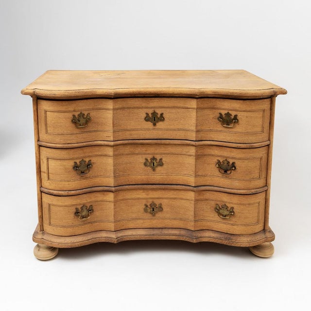 A three-drawer commode made of light softwood with a distinct serpentine front. The piece features a triple-curved shape...
