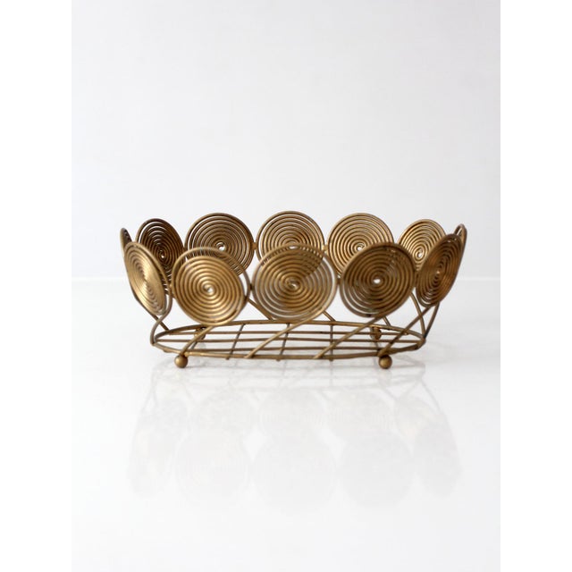 Vintage mid-century brass-plated wire fruit basket featuring sculptural spiral medallions and an open oval frame. The warm...