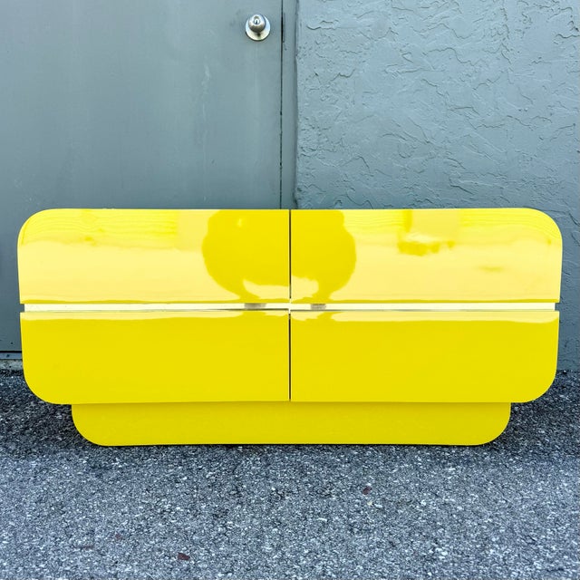 A bold postmodern chest of drawers featuring a vibrant high-gloss yellow laminate finish with sculptural waterfall edges...
