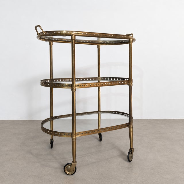 Vintage three-tier serving trolley featuring a brass frame and glass shelves. The oval structure is mounted on casters....