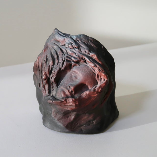 Striking handmade ceramic sculpture featuring a softly modeled human face emerging from an abstract, cocoon-like form. The...