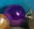 This oil painting features a still life composition of three onions and a vibrant purple ornament, set against a deep blue...