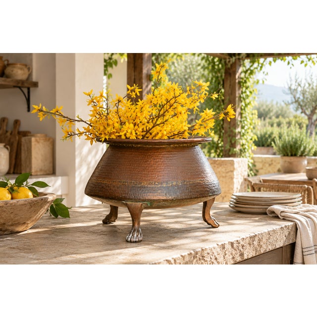 Hammered copper swells into a wide, flared bowl with a rolled rim, resting on three brass legs that end in carved paw...