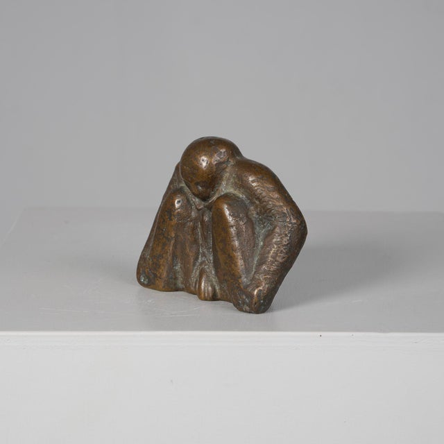 An expressive bronze sculpture depicting a crouching male figure in a compact, introspective pose. The body is reduced to...