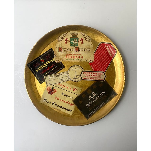 A round cocktail serving tray with gold leaf and decoupage collage of champagne and spirits labels, circa mid‑20th...