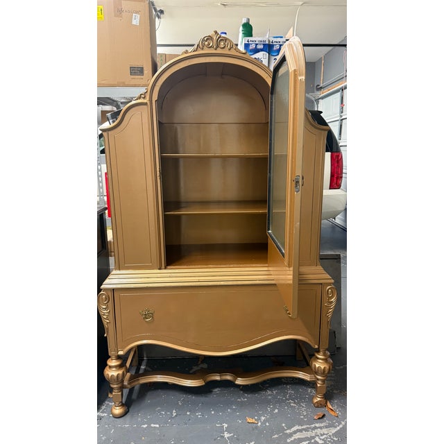 Vintage French Provincial–style gold display cabinet with an arched top, ornate carved details, and a frosted glass center...