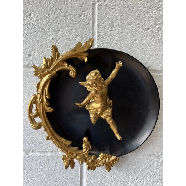 A striking pair of vintage Hollywood Regency wall plaques featuring sculptural gilt-metal cherubs against glossy black...