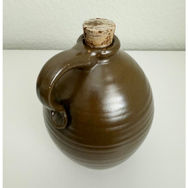 Lovely vintage or antique earthenware jug or bottle with a curved handle and cork stopper. Features a chocolate brown...