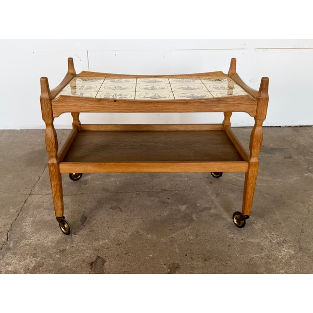 A solid oak two-tier bar trolley by Henning Kjærnulf, Denmark, 1960s. The upper tier is inset with twelve hand-painted...