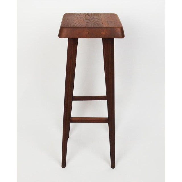 Solid elm bar stool in mahogany color. Square seat with rounded corners, legs connected by four bars. Model created in...