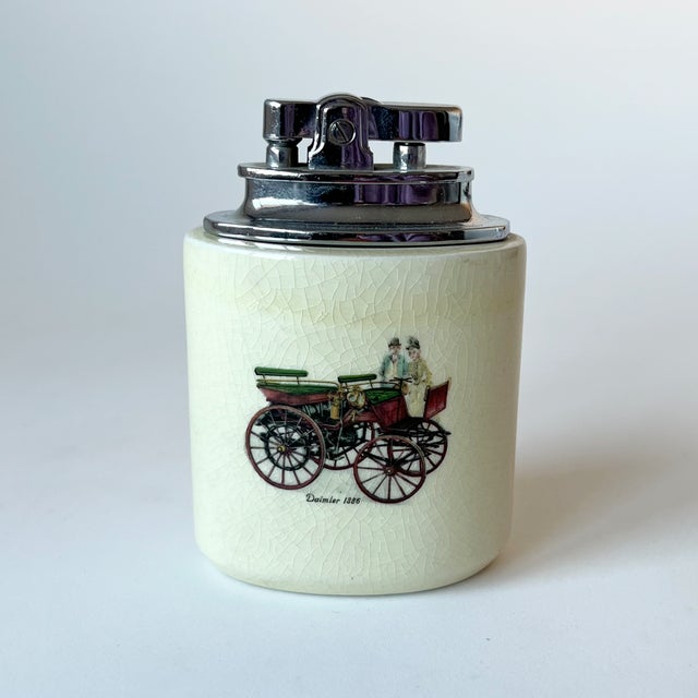 This table lighter features a cylindrical ceramic base with a glazed finish, bearing printed illustrations of a 'Daimler...
