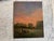 Atmospheric contemporary oil painting depicting two grazing cows in an open meadow at dusk. The composition is dominated...