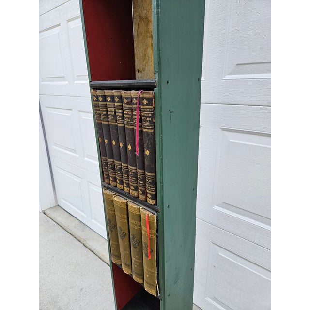 This handmade folk art slender bookcase features two central cabinets with faux book fronts, along with additional shelves...