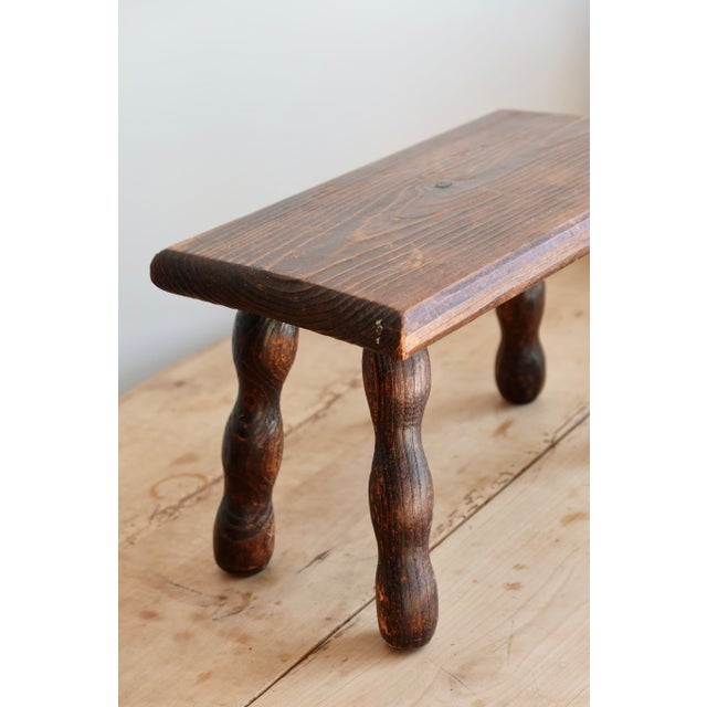 Low, rectangular stool in dark wood, resting on four turned legs with the typical curve of rustic furniture. Antique or...