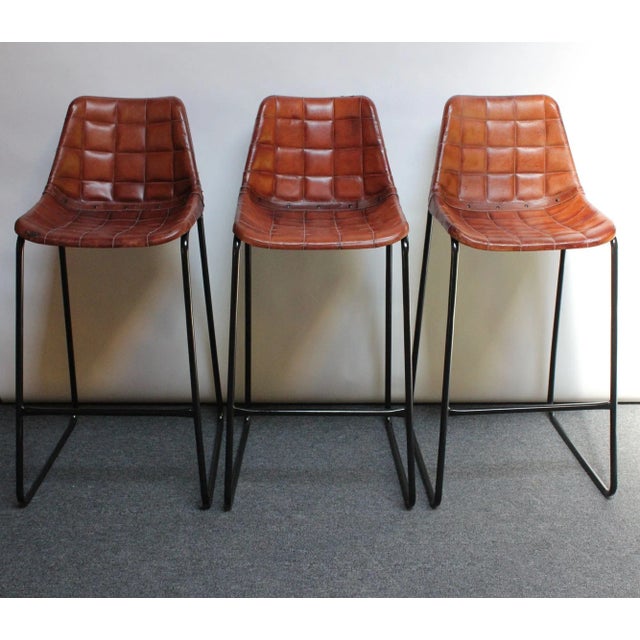 Set of three richly patinated barstools with leatherette seats supported by wrought iron and steel frames with footrests...