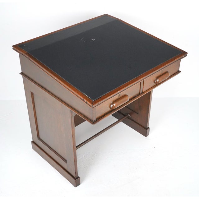 Antique slanted writing desk made of dark finished oak and a black leather top. It features two drawers and a footrest. No...