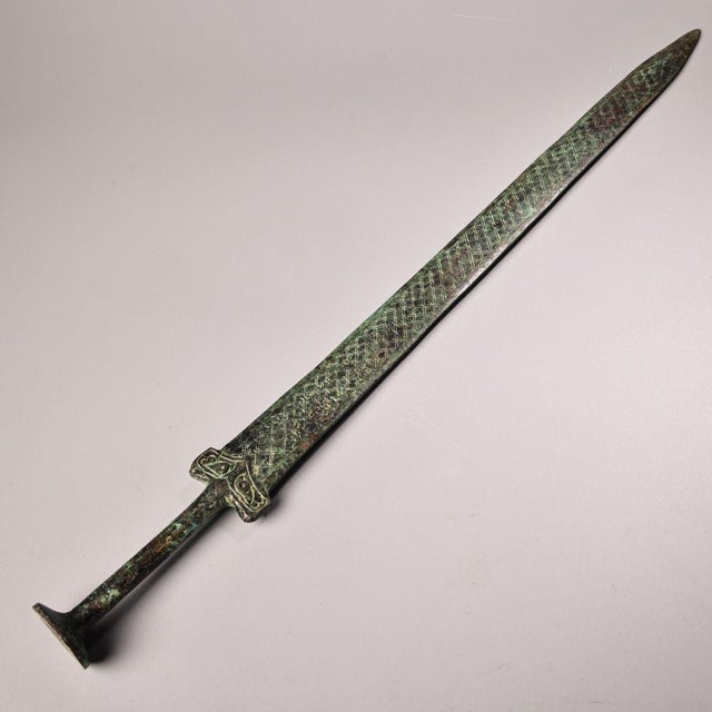 This is a cast bronze sword featuring a double-edged blade with a raised central ridge and a pointed tip. The blade is...