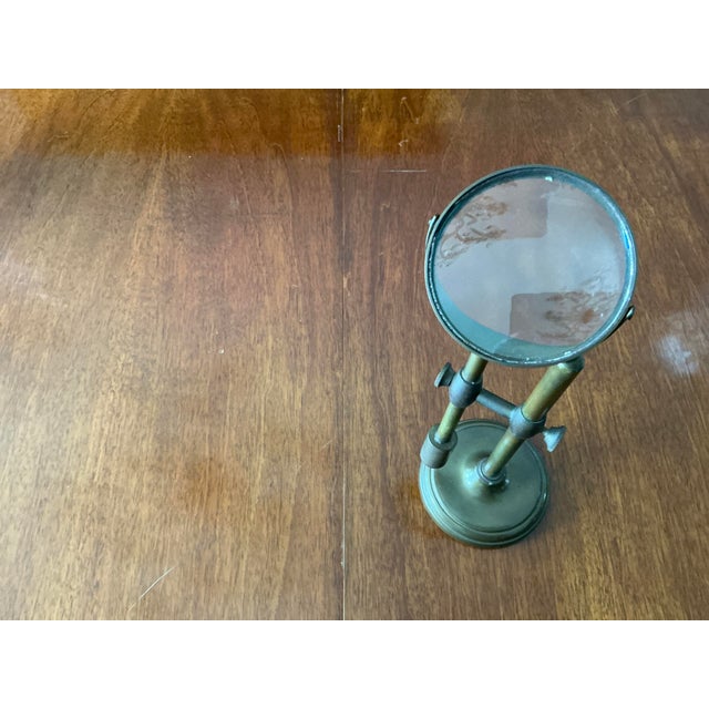 A brass classic design hands-free desktop magnifying glass on a large, circular weighted base. It features a crossbar with...