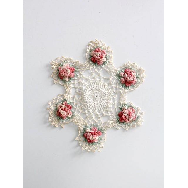 Vintage crocheted doily featuring raised floral rosettes arranged around an openwork center. Soft pink and cream tones...