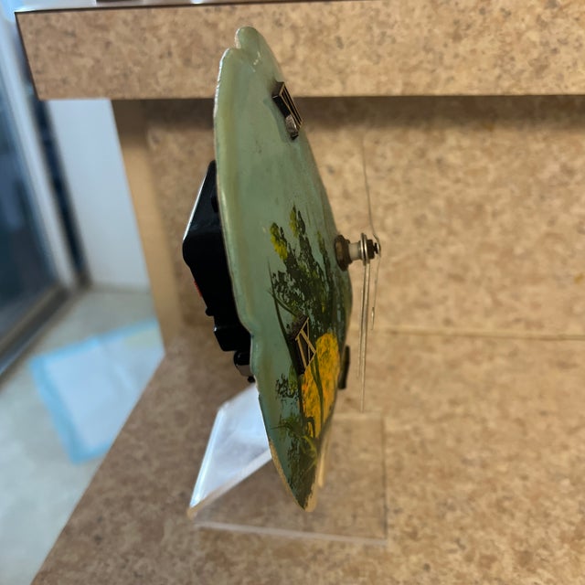 This item is a decorative desk clock crafted from a sand dollar, featuring a painted underwater scene with a fish....