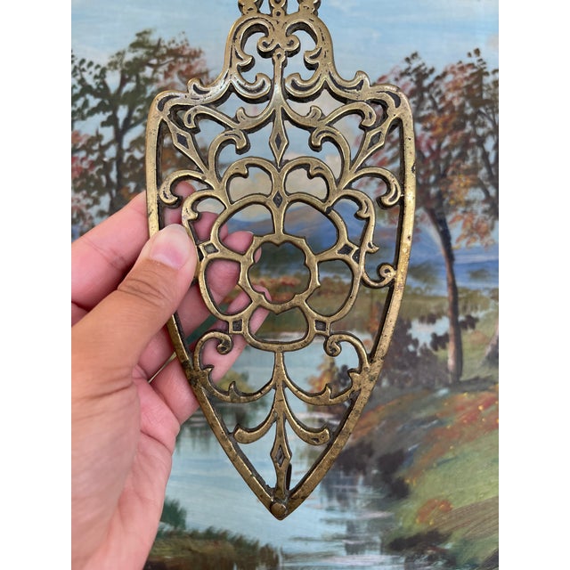 A w toned trivet featuring an ornate openwork design in the Victorian style. The trivet is shaped in a shield-like form...