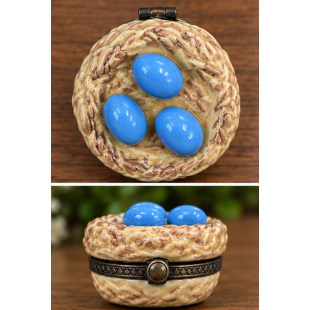 Charming hand-painted porcelain hinged trinket box designed as a bird’s nest holding three glossy blue eggs. The textured...