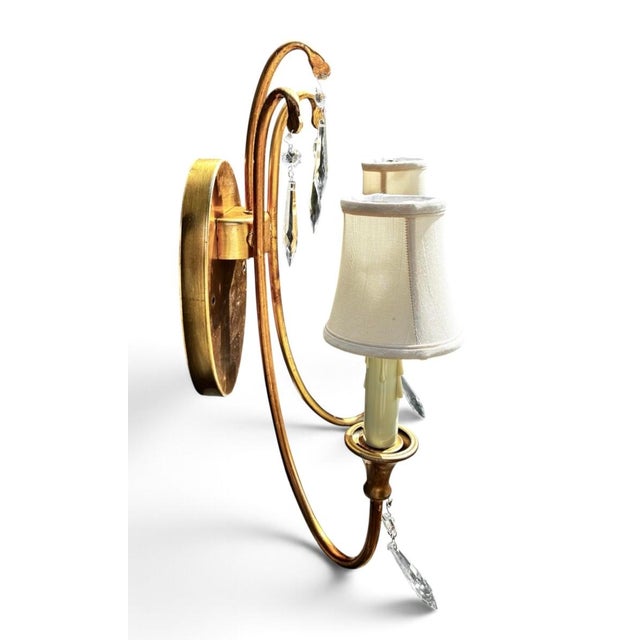 Elegant wall sconce has a gold leaf finish on a metal frame. This two-light fixture is adorned with hanging crystal prisms...