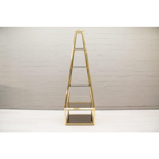 This shelving unit features a golden brass frame, smoked glass shelves, and a black-lacquered, removable base.