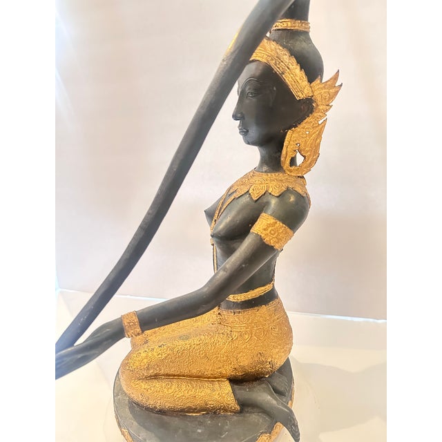 An elegant black and gold sculpture depicting a kneeling female figure in traditional Southeast Asian attire. The figure...
