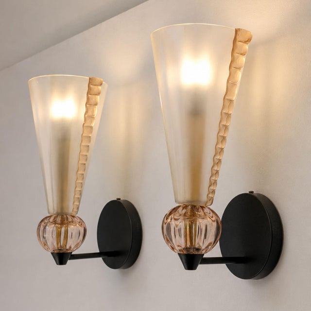 A pair of Mid-Century Modern wall lamps attributed to Barovier & Toso, featuring a black metal frame and pink Murano glass...